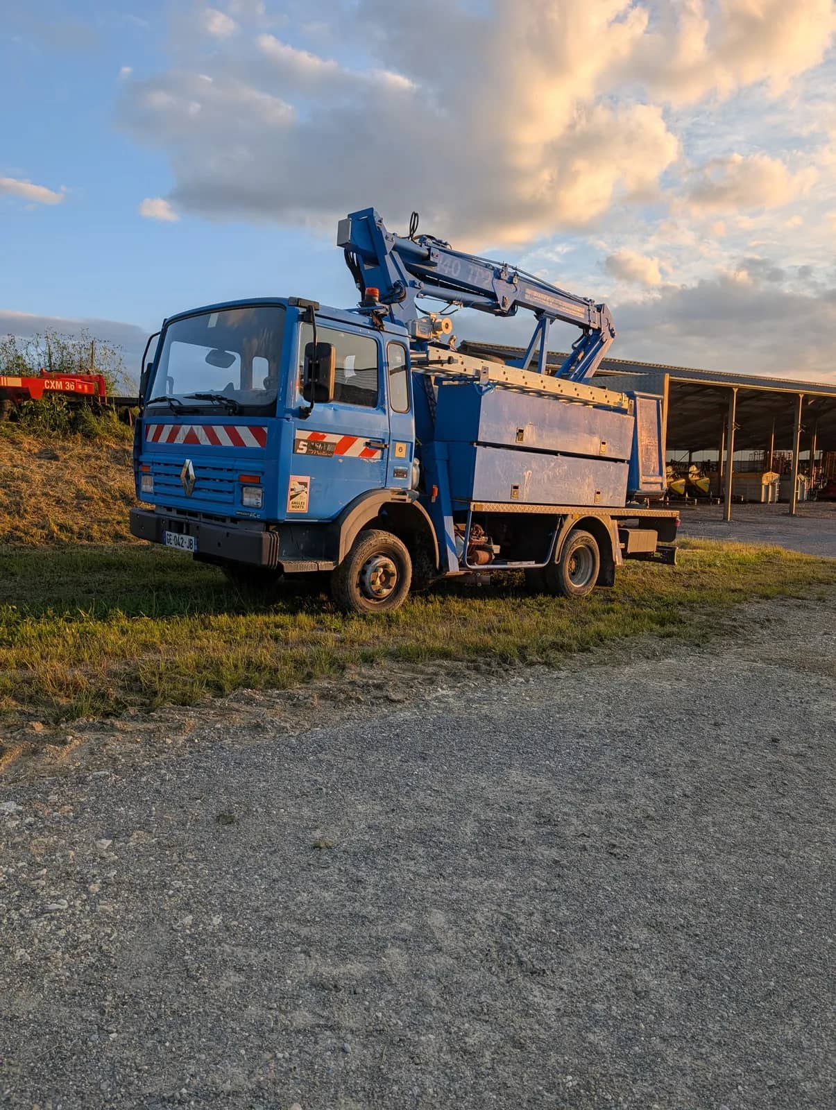 Camion bleu, nacelle au bras replié, garé sur un chemin de gravier devant un hangar