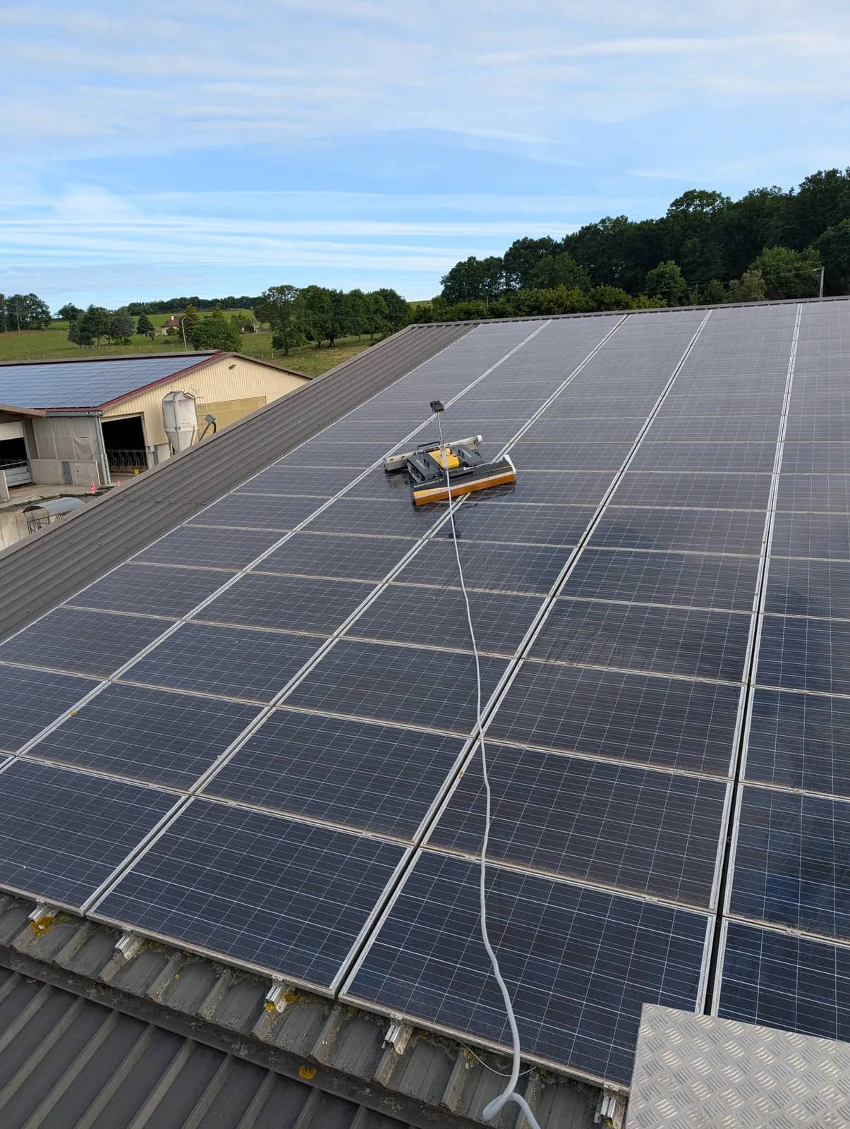 Robot SolarCleano F1 et son tuyau sur la toiture de panneaux d'un bâtiment agricole, un hangar voisin