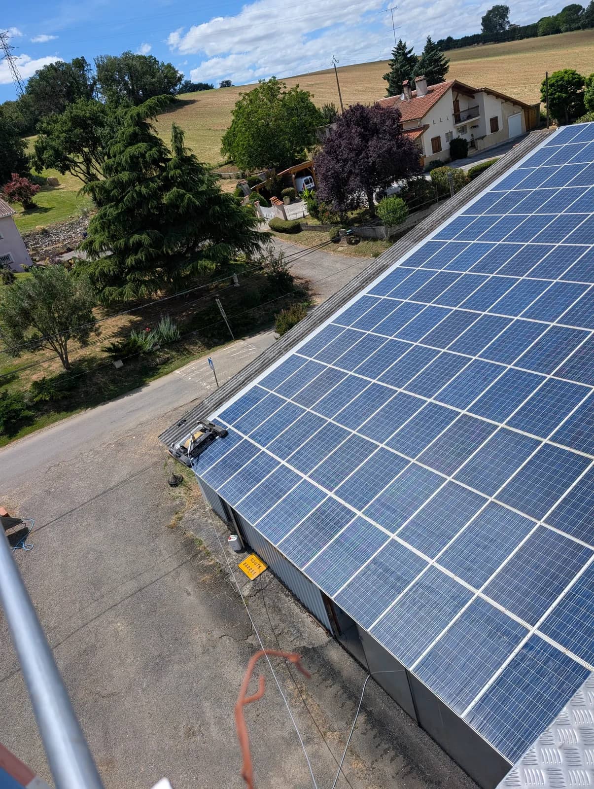 Toiture photovoltaïque en pente sur un bâtiment agricole en métal, un robot en bas de rangée, champs et une maison à l'arrière-plan, vue depuis une nacelle
