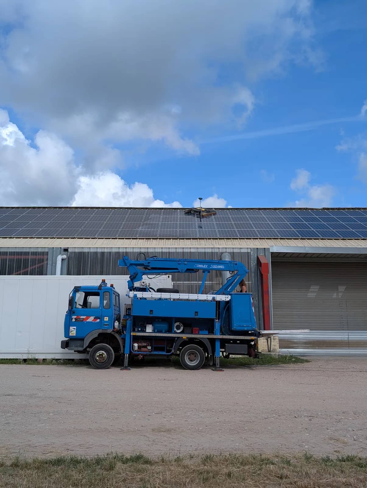 Camion nacelle bleu, stabilisateurs sortis, devant un bâtiment dont le toit est couvert de panneaux, un robot sur la toiture