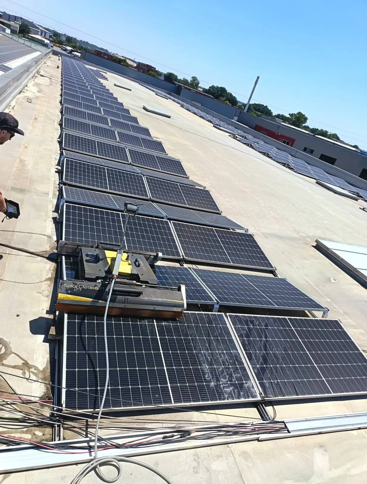 Toit plat, rangées de panneaux dont plusieurs sont ternis, un robot SolarCleano, une personne au bord du toit et des câbles au premier plan